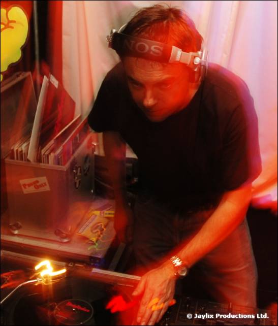 A DJ in headphones leans over the decks beside a box of records
