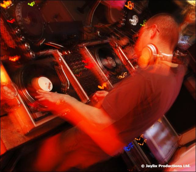 A DJ in headphones mixes at Pioneer decks in a dark, orange-lit booth