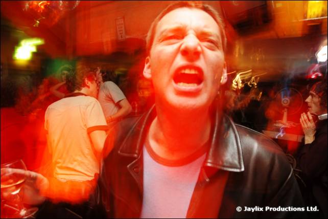 A man in a leather jacket shouts at the camera in a crowded bar