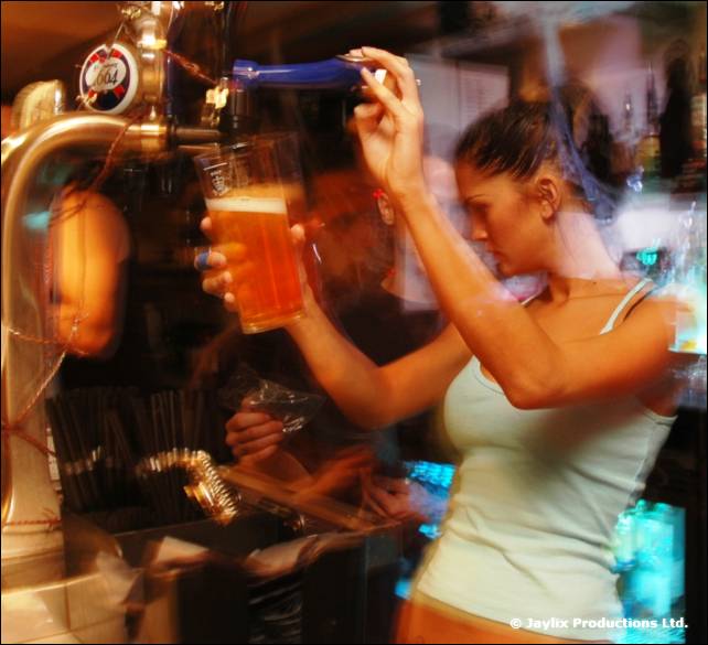 A woman in a white vest raises a pint of beer at a bar with a blurred tap beside her