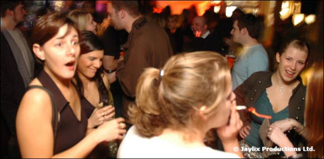 Three women stand talking in a busy bar, one holding a cigarette