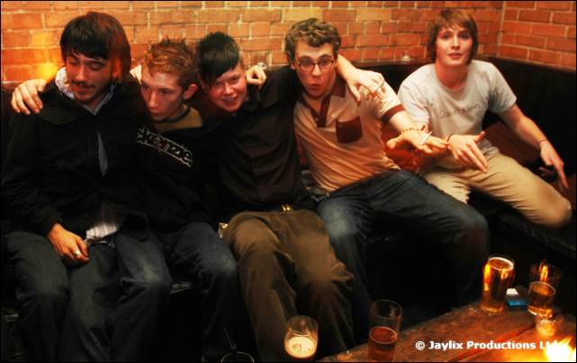Five men sit close together on a sofa against a brick wall with drinks on the table