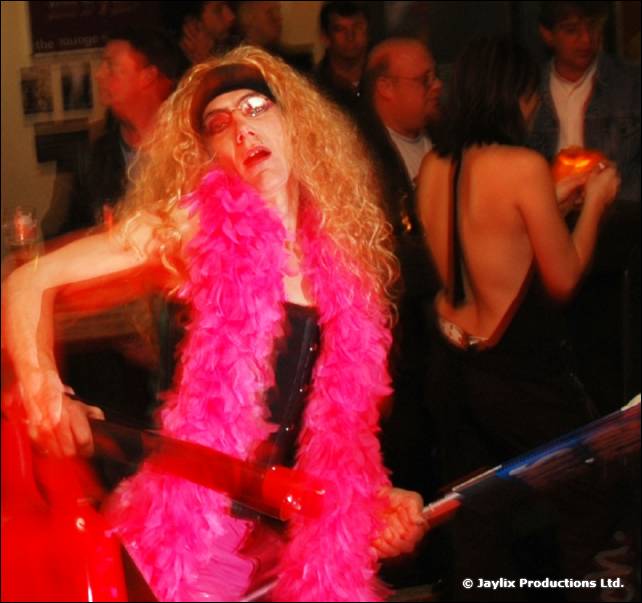 A person with a long curly blonde wig and pink feather boa dances with a red prop