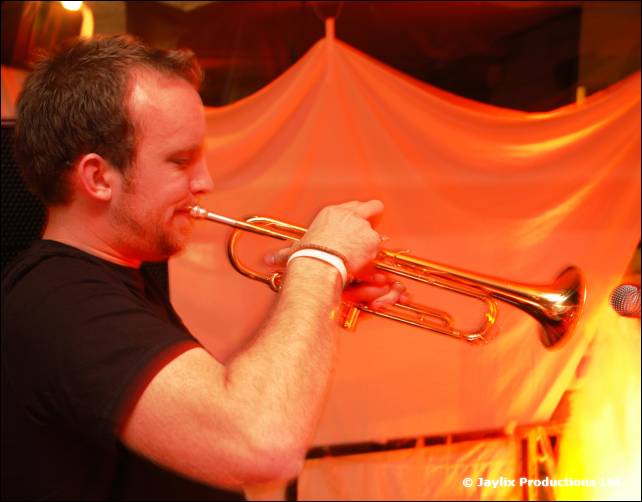 A man in a black T-shirt plays a trumpet in front of an orange curtain