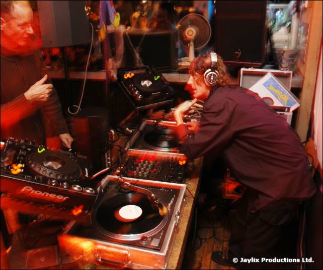 A DJ in headphones plays at Pioneer decks while another man looks on from the left