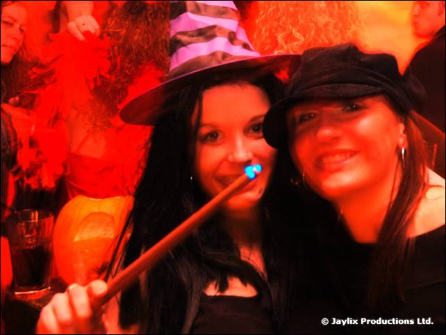 Two women in dark hats pose close together, one in a witch hat holding a wand with a blue light