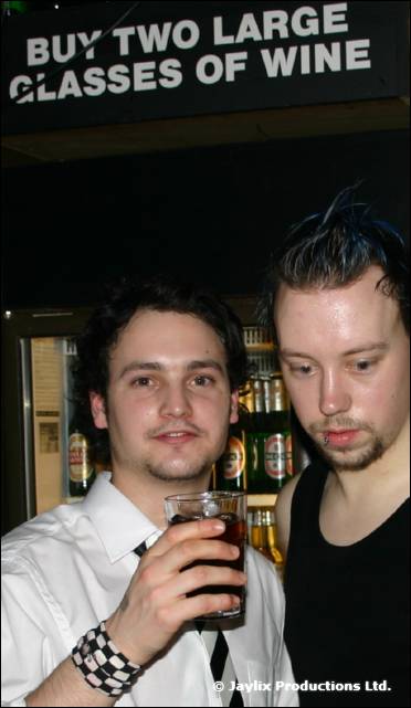Two men stand at a bar beneath a sign reading 'Buy two large glasses of wine', one holding a drink