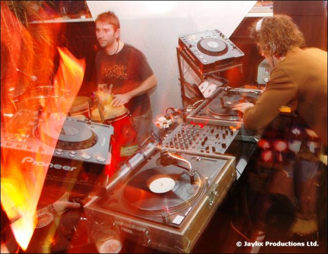 A person plays drums beside a DJ at Pioneer decks, with fire in the foreground
