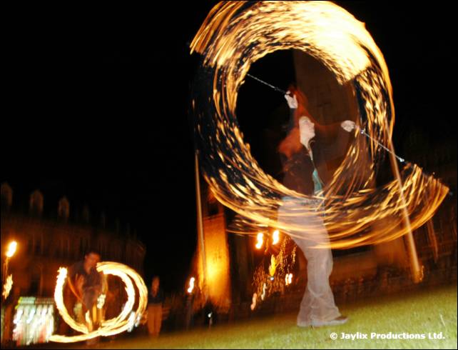 A woman spins flaming poi, tracing large rings of fire, on a dark lawn with a second fire spinner at the left
