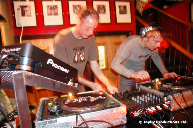 Two DJs in grey T-shirts work at Pioneer decks in front of framed pictures on a red wall