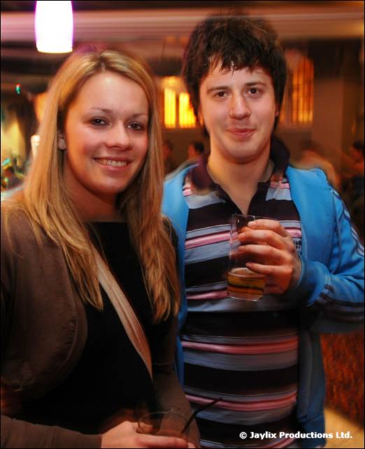 A smiling woman with long blonde hair stands beside a man in a striped top and blue jacket, holding a drink