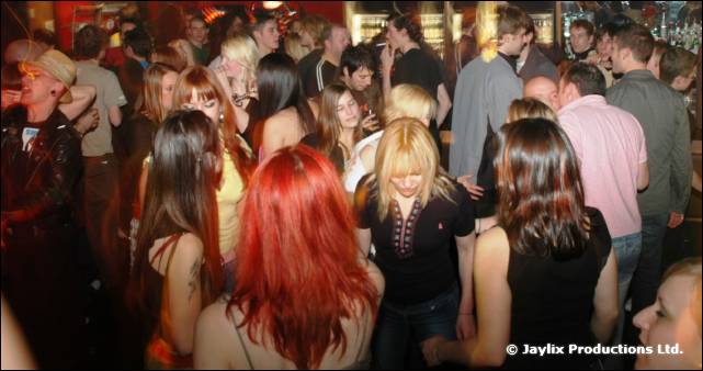 A packed crowd dances in a bar, with a red-haired woman in the foreground