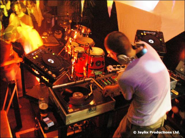 A DJ in a white T-shirt mixes beside drums, with an orange glow from flames behind