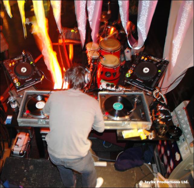 A DJ in a light jumper plays at two turntables seen from above, with drums and flames behind