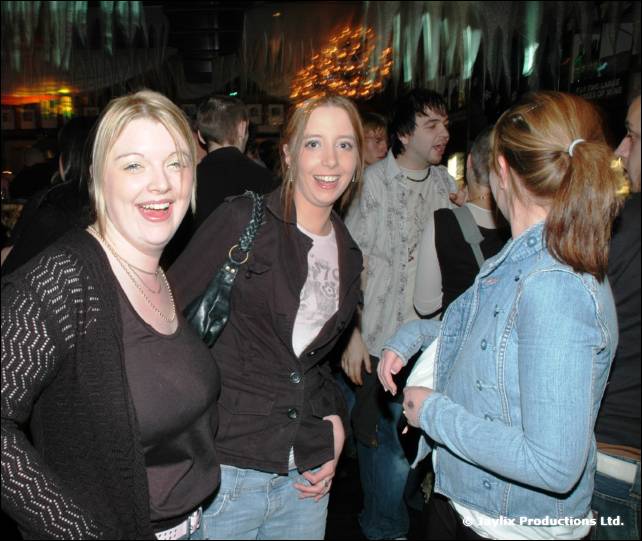 Three women smile at the camera in a busy bar under orange lights
