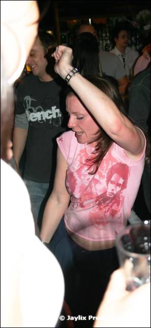 A woman in a pink T-shirt dances with one arm raised in a crowded bar