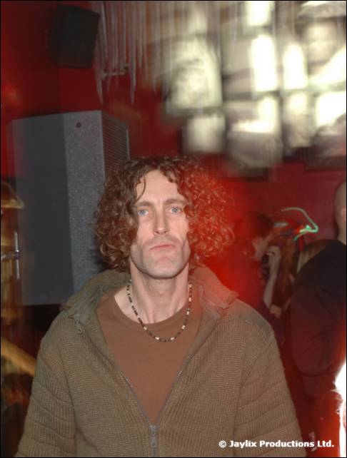 A man with curly hair in a green jacket stands unsmiling in a red-lit room