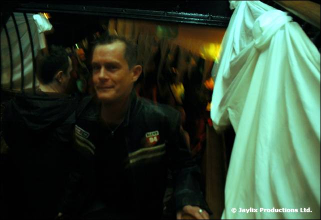 A man in a dark jacket smiles in a dark bar, with a pale hooded figure beside him