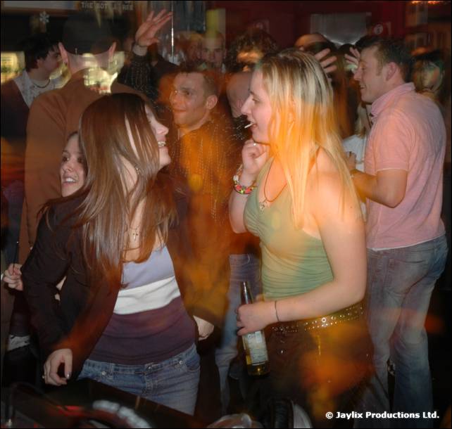 A woman with long hair dances in a crowd beside a woman holding a bottle and a cigarette