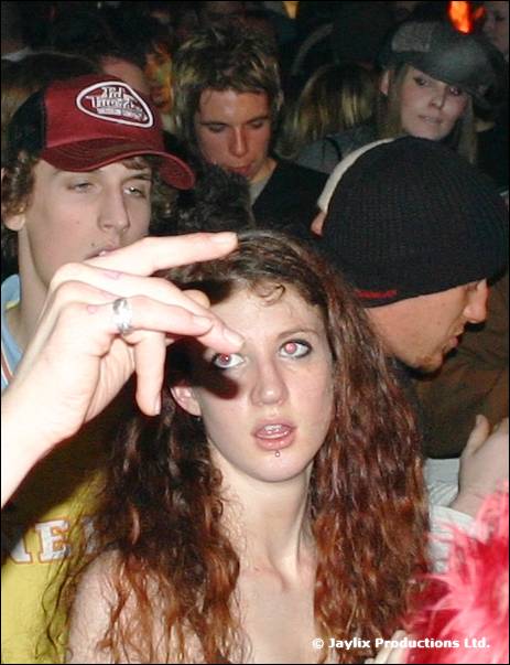 A woman with long curly hair looks at the camera among a crowd, with a hand held up in front