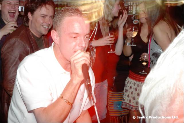 A man sings into a microphone in a white shirt, with people holding glasses behind him