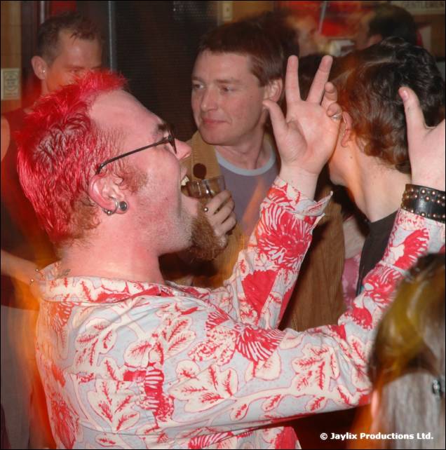 A man with red spiky hair and glasses sings with one hand raised, in a crowded bar