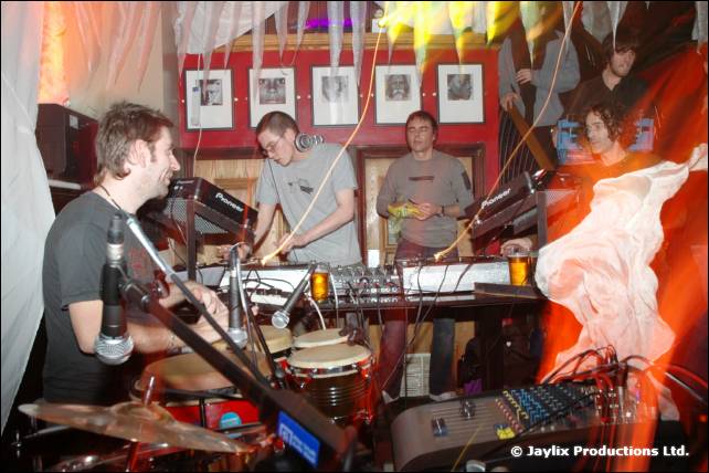 A band plays keyboards and drums in front of framed pictures on a red wall