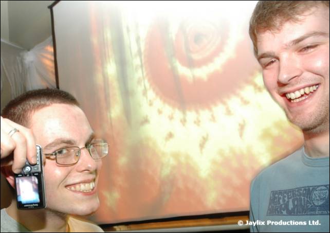 Two smiling men in glasses, one holding a mobile phone, stand in front of a projection screen