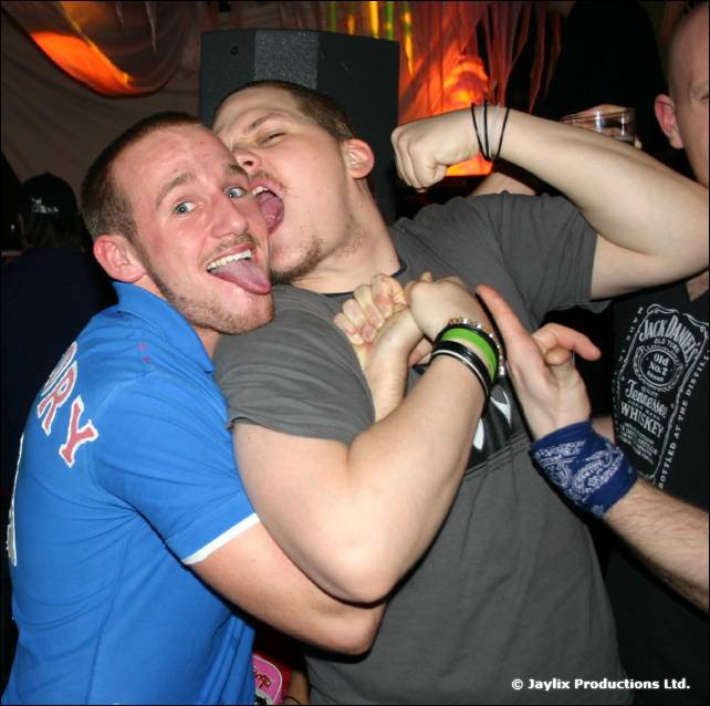 Two men grapple together, one with his tongue out, in a bar