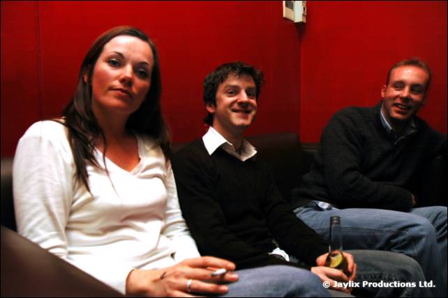 A woman and two men sit on a red sofa, the woman and one man holding cigarettes