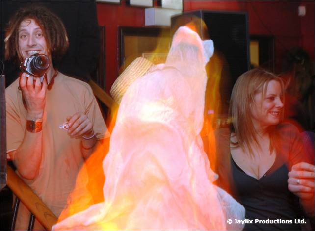 A man with a camera stands beside a blurred glowing pale figure, with a woman on the right