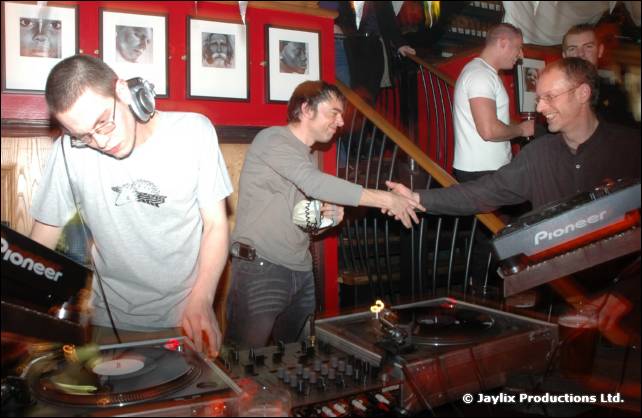 A DJ in a white T-shirt mixes while two men shake hands behind him, with Pioneer equipment in front
