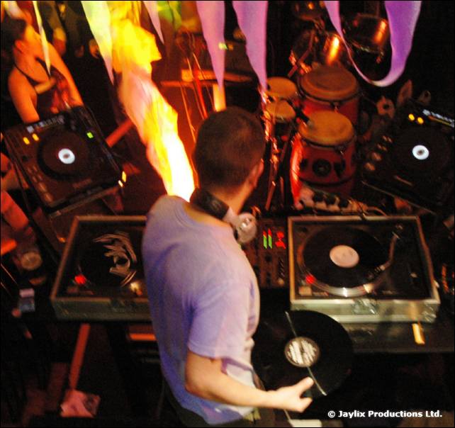 A DJ in a light top plays at two turntables seen from above, with drums and flames behind