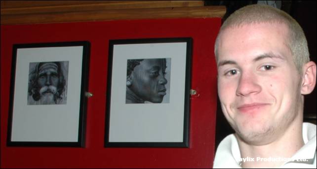 A smiling man with short hair stands beside two framed black and white portraits on a red wall