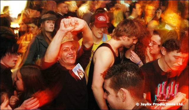 A crowd of people dance in a bar, one man in a straw hat raising his fist