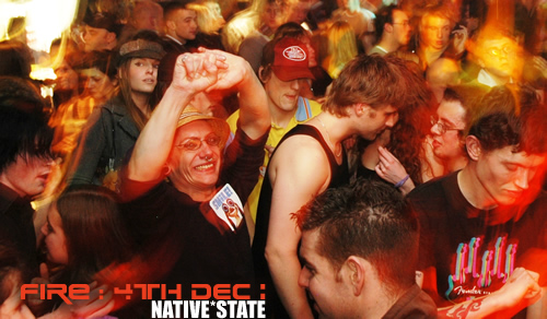 A crowd dancing in a bar under a banner reading FIRE: 4th Dec: Native State, a man in a hat smiling at the front