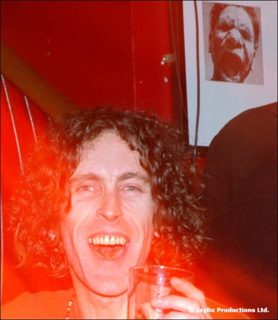 A man with curly hair laughs while holding a glass, with a framed portrait on the wall behind him