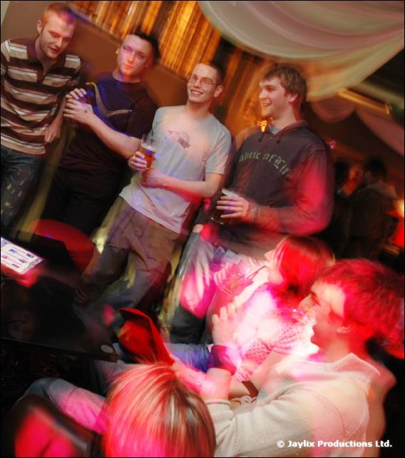 Five men stand in a group in a bar holding drinks, with people sitting in the foreground