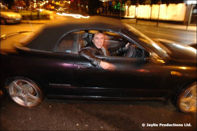 A man smiles from the driver's seat of a dark convertible car on a night street