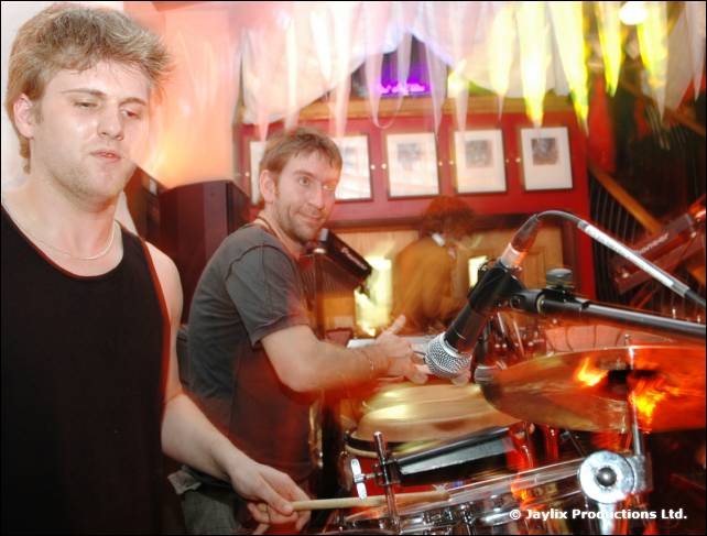 Two men play drums and percussion beside microphones, with framed pictures on the wall behind