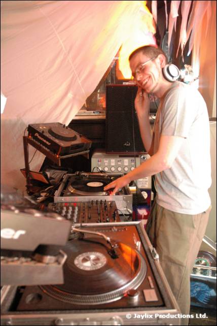 A DJ in a grey t-shirt and headphones leans his head on his hand beside turntables and a mixer