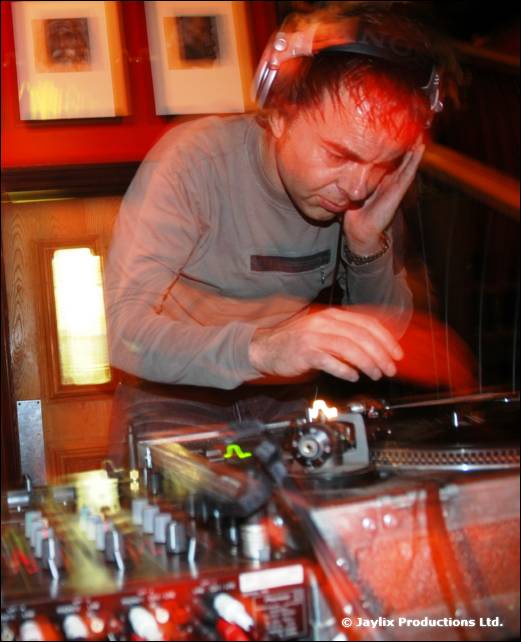 A DJ in a grey top and headphones rests his head on one hand while mixing at decks, motion blurred
