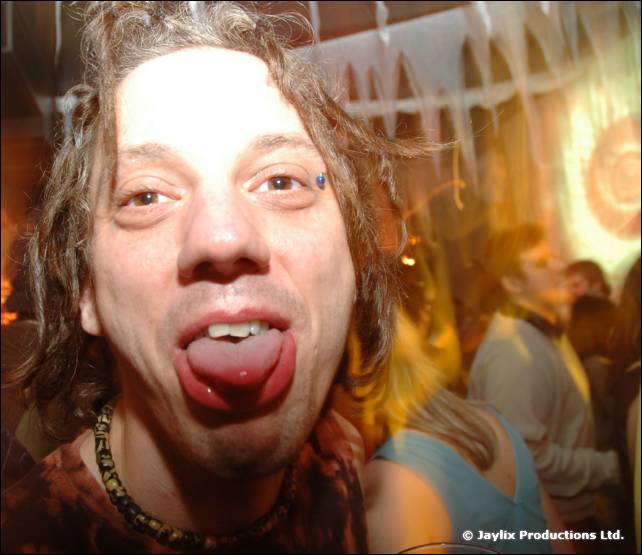 Close-up of a person with messy hair sticking their tongue out at the camera in a crowded room