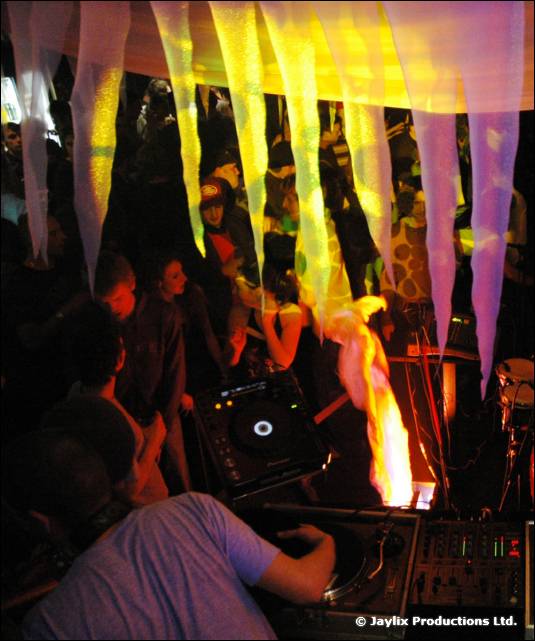 A person at CDJ decks under hanging white and yellow streamers, with a flame and dancers behind