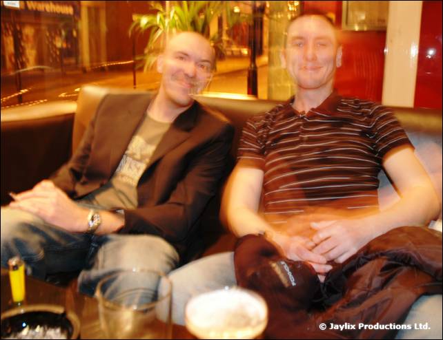 Two smiling men sit on a leather sofa, one in a dark blazer, one in a striped polo shirt, with drinks in front