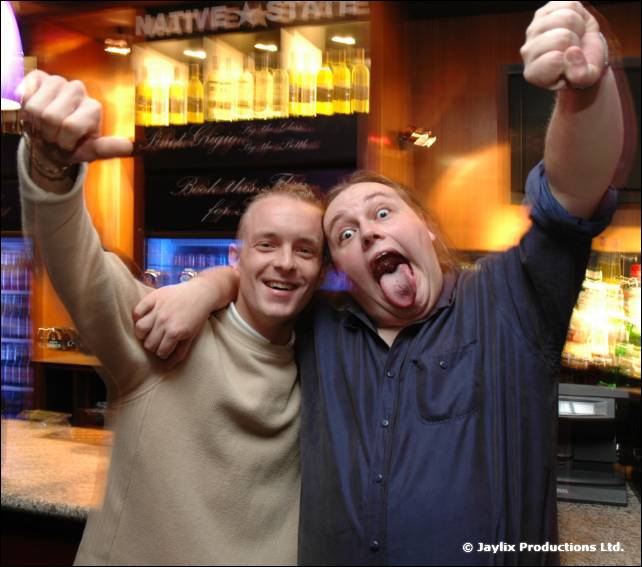 Two men with arms raised cheer at the camera, one sticking his tongue out, in front of a Native State bar