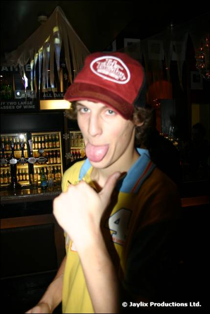 A person in a red cap and yellow top sticks their tongue out and gives a thumbs up in a bar