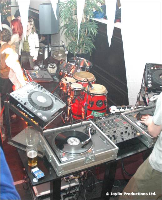 DJ decks with turntables and a mixer beside a set of red congas, with people dancing behind