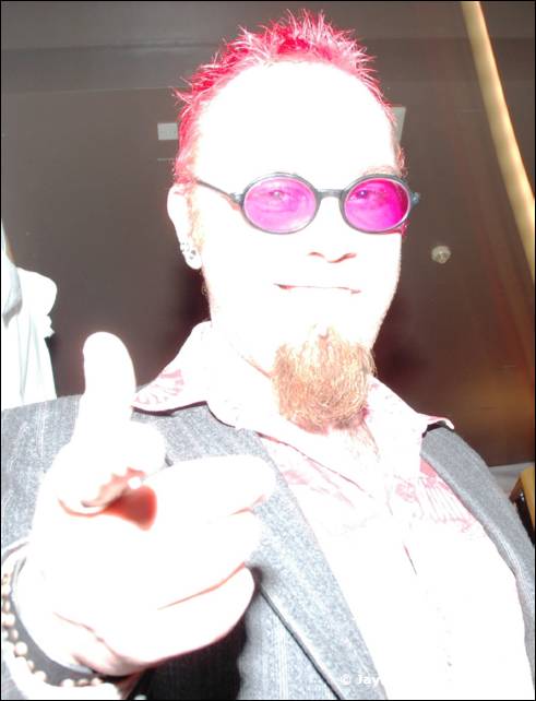 A person with red spiky hair, pink round glasses and a goatee points at the camera