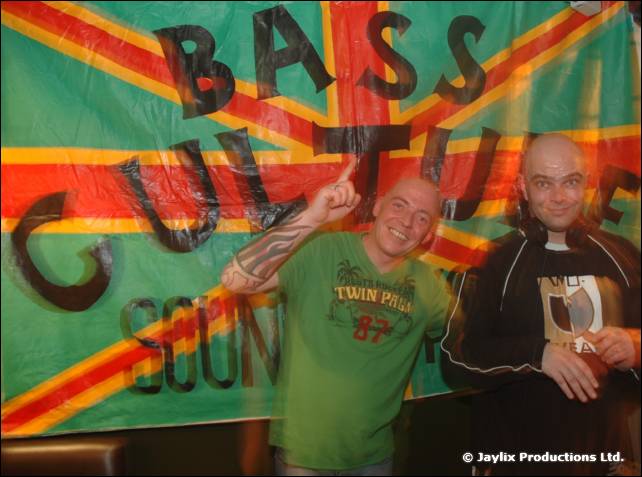 Two bald men pose in front of a green, red and yellow Bass Culture Sound banner